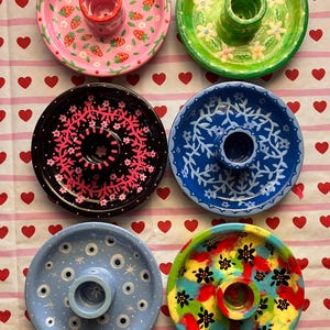 May include: Six colorful, hand-painted candle holders. The candle holders are round with a central cup for the candle. Designs include strawberries, flowers, and abstract patterns. The colors are pink, green, black, blue, light blue, and multi-colored.