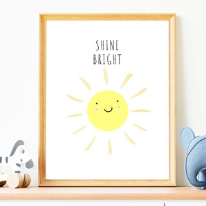 以下が含まれることがあります： 「SHINE BRIGHT」の文字と、笑顔の黄色い太陽のイラストが描かれたフレーム入りプリント。太陽は笑顔で、薄い黄色の光線があります。フレームは明るい茶色の木製です。木製のシマウマのおもちゃと青い象のおもちゃが棚に置かれています。