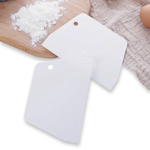 May include: Two white plastic cake decorating tools with a hole in the top of each. The tools are on a wooden surface with a white cloth and a bowl of white powder in the background.