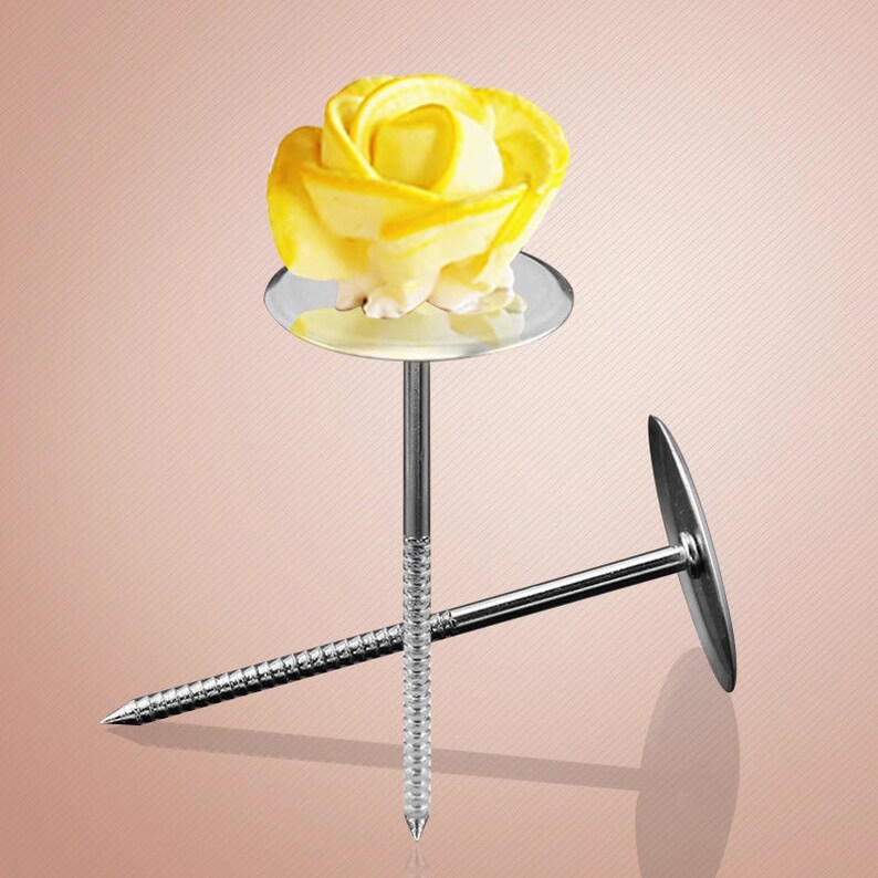 May include: Two silver cake decorating tools with round bases and long, thin, pointed stems. One tool has a yellow buttercream rose on top of the base.
