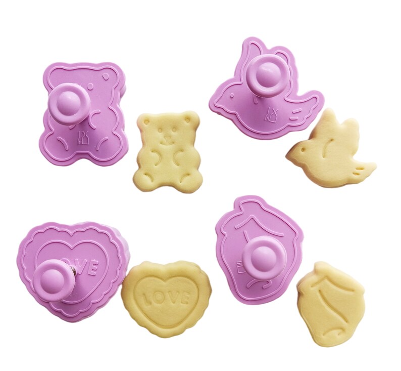 May include: Set of eight pink plastic cookie cutters in various shapes, including a bear, a bird, a heart, and a house. The cutters are shown with matching cookies.