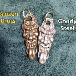 Puede incluir: Dos llaveros con un diseño de rostro grotesco detallado. Un llavero es de latón y es de color dorado. El otro llavero es de acero y es de color plateado. El texto "Brazen Brass" está sobre el llavero de latón y el texto "Gnarly Steel" está sobre el llavero de acero.