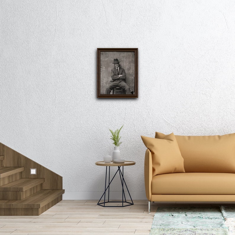 May include: A black and white photograph of a man wearing a hat and suit, sitting on a stool. The photo is framed in a dark wood frame and is hanging on a white wall.