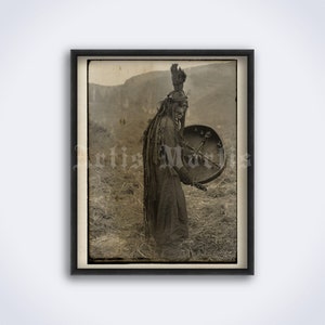 May include: A black and white photograph of a person wearing a headdress and holding a drum. The person is standing in a field of grass.