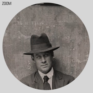 May include: A black and white photo of a man wearing a dark suit and a fedora. The man is looking directly at the camera. The photo is taken in a studio setting and the background is a plain wall. The text "S.H. ELLIS" is visible on the wall.