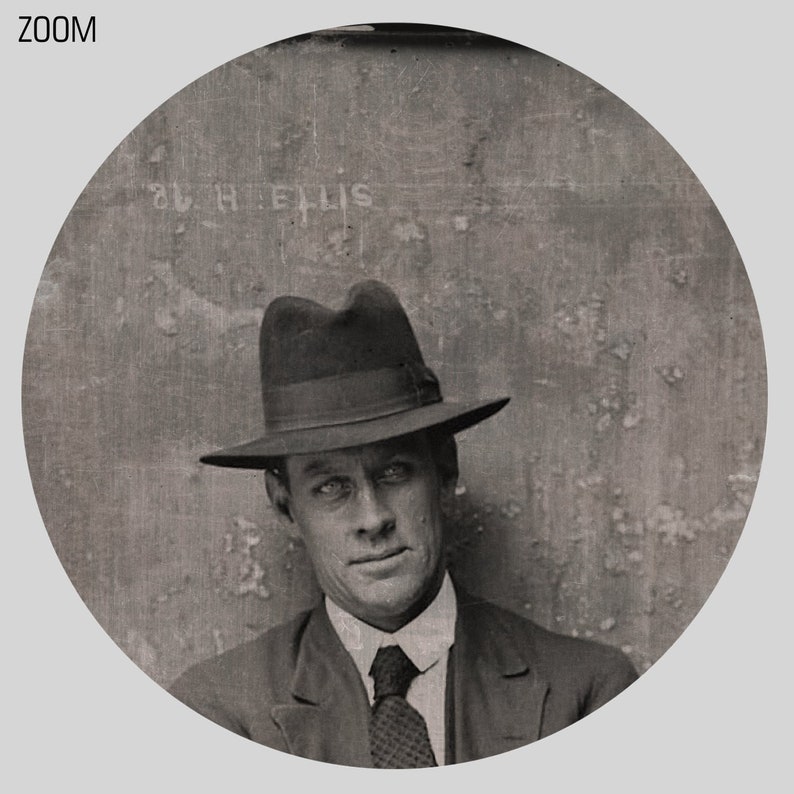 May include: A black and white photo of a man wearing a dark suit and a fedora. The man is looking directly at the camera. The photo is taken in a studio setting and the background is a plain wall. The text "S.H. ELLIS" is visible on the wall.