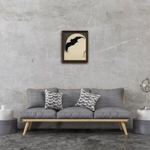 May include: Framed artwork featuring a black bat silhouette against a cream-colored moon. The bat is in flight, with a dark brown frame. The artwork is displayed above a gray sofa with decorative pillows.