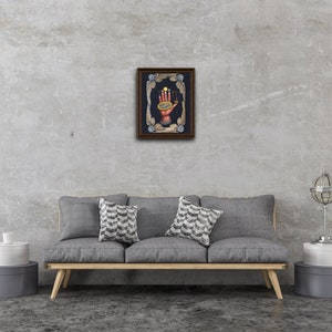 May include: A framed artwork depicting a hand holding a celestial sphere with planets and stars. The hand is surrounded by a decorative frame with wings and celestial symbols. The background is a dark blue.