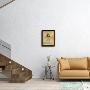 May include: A vintage-style wanted poster with a brown wood frame. The poster features a black and white illustration of a man with a hat and a gun. The text on the poster reads "WANTED", "Alphonse Gabriel Capone", "Scarface", "&pound;10,000 REWARD", and other details about the man.