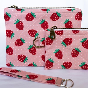 Puede incluir: Dos estuches de tela acolchada y una correa de muñeca a juego con un patrón repetitivo de fresas rojas con hojas verdes sobre un fondo rosa claro con lunares blancos. Cada estuche tiene un cierre de cremallera de metal color latón.