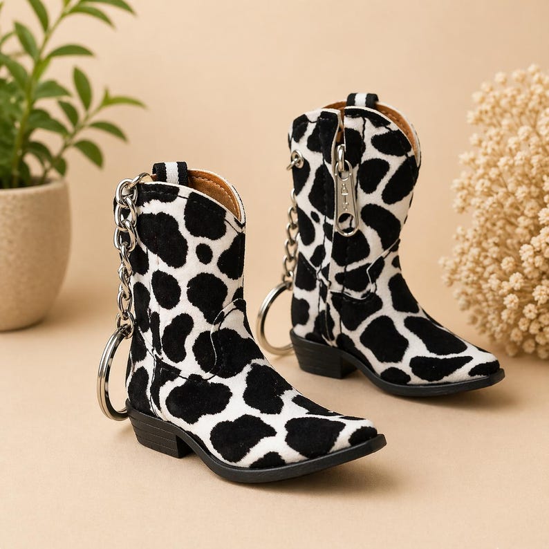 Peut inclure: Deux mini-bottes de cowboy avec un motif de peau de vache noir et blanc. Chaque botte a une cha&icirc;ne et un porte-cl&eacute;s argent&eacute;s. Les bottes ont une fermeture &eacute;clair et un petit talon. Les bottes sont sur un fond neutre.
