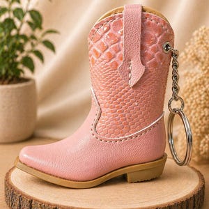 May include: A miniature pink cowboy boot keychain with a silver key ring. The boot features a textured, snake-like pattern on the upper shaft and a smooth leather finish on the lower part. The boot is set on a small wooden slice.