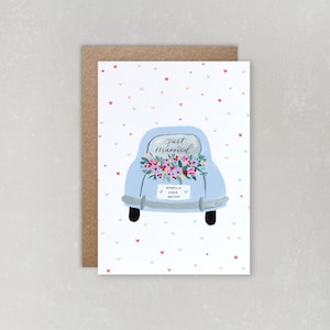 Puede incluir: Un coche azul claro con una corona de flores en el cap&oacute; y un letrero que dice "Just Married" y "Happily Ever After". El coche est&aacute; sobre un fondo blanco con un patr&oacute;n de peque&ntilde;os corazones rojos, rosas y azules.