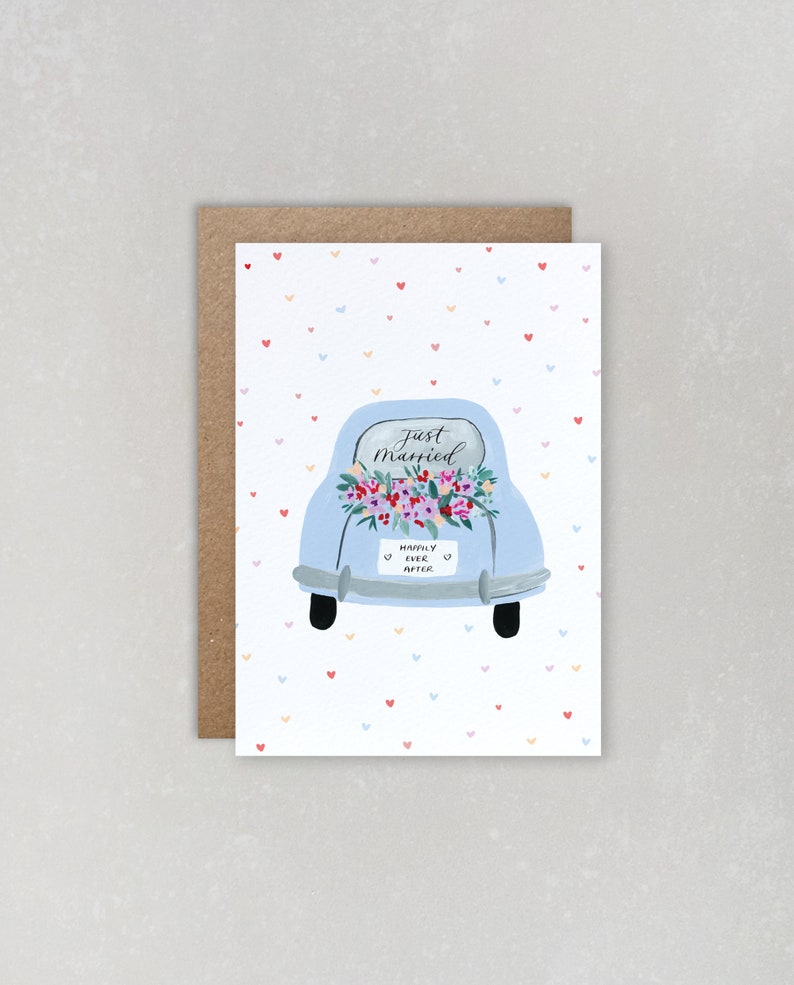 Puede incluir: Un coche azul claro con una corona de flores en el cap&oacute; y un letrero que dice "Just Married" y "Happily Ever After". El coche est&aacute; sobre un fondo blanco con un patr&oacute;n de peque&ntilde;os corazones rojos, rosas y azules.