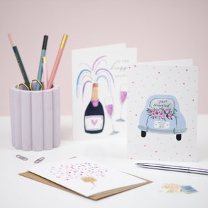 Puede incluir: Cuatro tarjetas de felicitaci&oacute;n de boda con diferentes dise&ntilde;os. Una tarjeta presenta una botella de champ&aacute;n con un arco&iacute;ris de confeti, otra tarjeta tiene un coche con las palabras "Just Married" y "Happily Ever After", una tercera tarjeta tiene una botella de champ&aacute;n con confeti, y la &uacute;ltima tarjeta tiene una botella de champ&aacute;n con confeti.