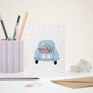 Puede incluir: Una tarjeta blanca con un coche azul decorado con flores y el texto "Just Married" y "Happily Ever After". El fondo est&aacute; cubierto de peque&ntilde;os corazones rosas, rojos y azules.