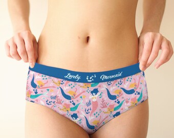 May include: Pink underwear with a mermaid pattern. The design features mermaids, coral, and seaweed in shades of blue, yellow, and pink. The waistband is blue with the words "Lovely Mermaid".