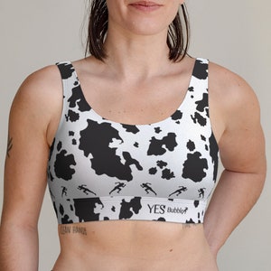 Pode incluir: Sutiã desportivo branco e preto com um design de estampa de vaca. O sutiã apresenta um padrão repetido de figuras de corrida ao longo da borda inferior e o texto "YES Bubble".