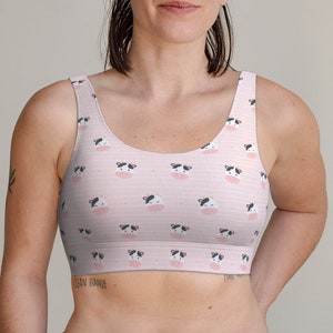 May include: A light pink sports bra with a repeating pattern of cartoon cow faces. The bra has a scoop neck and a racerback design. The cow faces are black and white with pink noses, set against a striped background.
