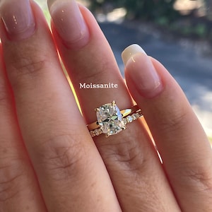 May include: A gold ring set featuring a central cushion-cut Moissanite stone held by four prongs on a plain band, paired with a matching gold eternity band encrusted with small round white stones, displaying the text Moissanite above the ring.