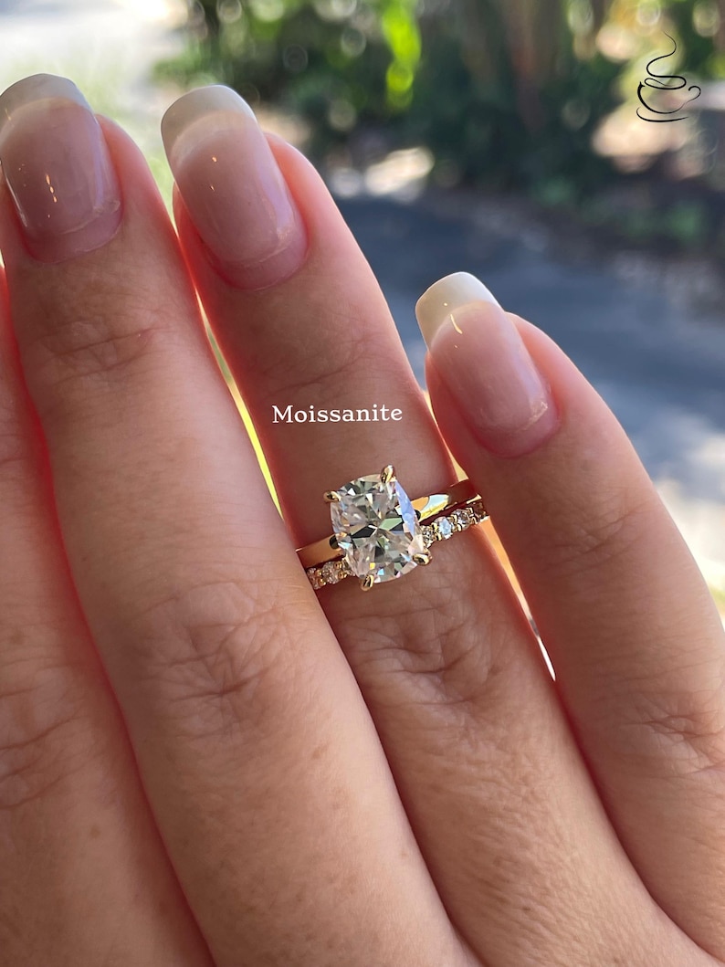 May include: A gold ring set featuring a central cushion-cut Moissanite stone held by four prongs on a plain band, paired with a matching gold eternity band encrusted with small round white stones, displaying the text Moissanite above the ring.