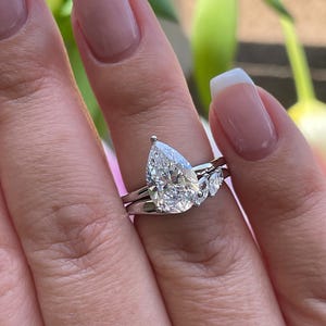 Könnte beinhalten: Ein birnenförmiger Diamant-Verlobungsring mit passendem Band. Der Ring hat einen großen, klaren birnenförmigen Diamanten als Mittelstein, flankiert von zwei kleineren Diamanten. Das Band ist dünn und silberfarben poliert.