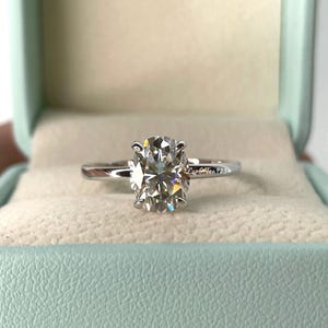 May include: A silver ring with a large, oval-shaped diamond in a solitaire setting. The ring is in a light blue ring box with the text "Cup Of Tea 725" on the side.