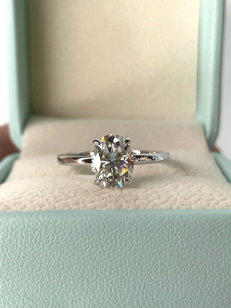 May include: A silver ring with a large, oval-shaped diamond in a solitaire setting. The ring is in a light blue ring box with the text "Cup Of Tea 725" on the side.
