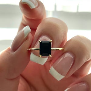 Könnte beinhalten: Ein goldener Ring mit einem rechteckigen, schwarzen Edelstein. Der Ring wird von einer Hand mit manikürten Nägeln gehalten. Der Text "CupOfTea325" ist auf dem Bild sichtbar.