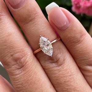 K&ouml;nnte beinhalten: Ein ros&eacute;goldener Ring mit einem Diamanten im Marquiseschliff. Der Diamant ist mit vier Krappen gefasst und funkelt hell. Der Ring befindet sich an einem Finger, mit einem verschwommenen Hintergrund aus gr&uuml;nem Laub und rosa Bl&uuml;ten.
