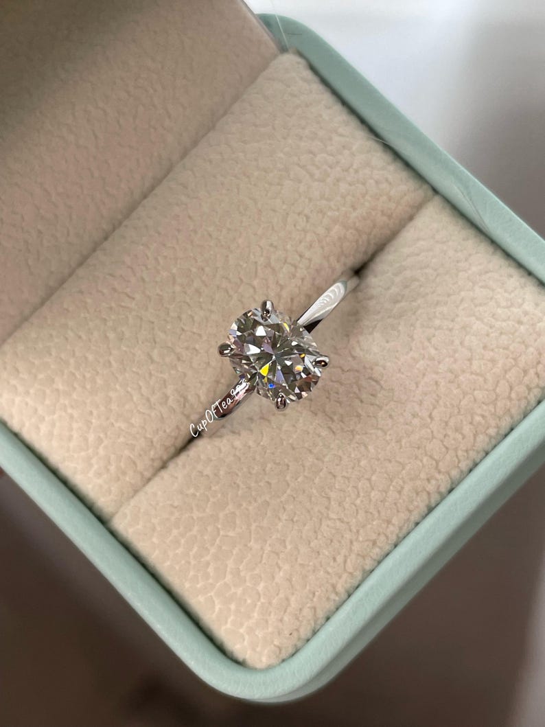 May include: A silver ring with a large oval-shaped diamond in a ring box. The ring is in a light blue box with a white interior.