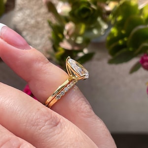 May include: A gold pear-shaped engagement ring with a pave band. The ring is set with a large, clear pear-shaped diamond. The band is set with smaller diamonds. The ring is shown on a finger.