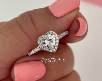 May include: A silver ring with a heart-shaped clear stone surrounded by smaller stones. The ring is on a finger with pink nail polish.