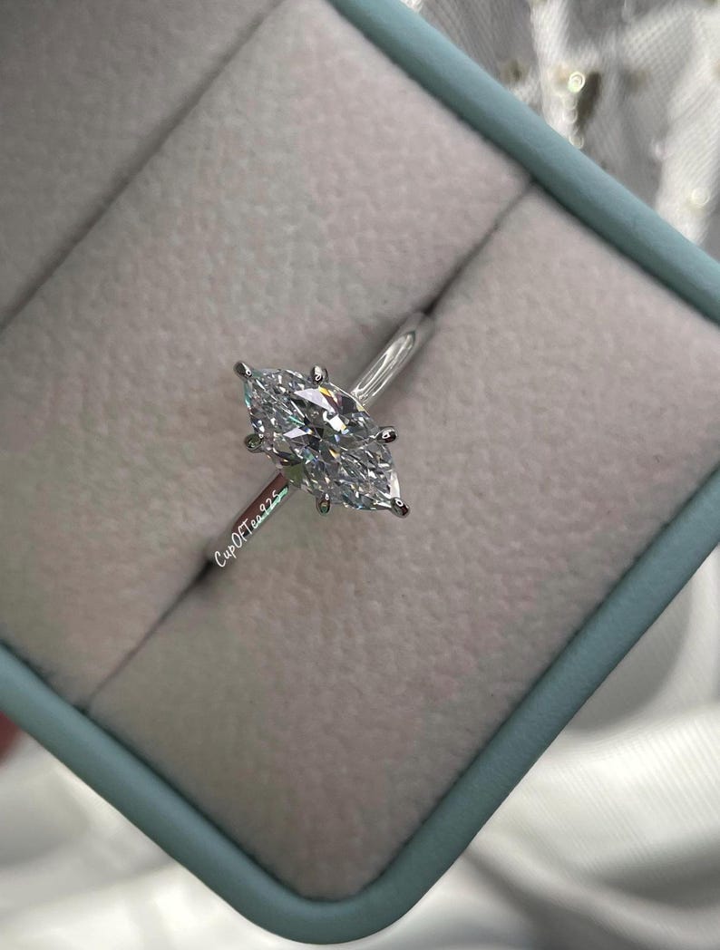 K&ouml;nnte beinhalten: Ein silberner Ring mit einem Marquise-Diamanten in einer hellblauen Ringbox. Die Ringbox hat den Text "CupOfTears" an der Seite.
