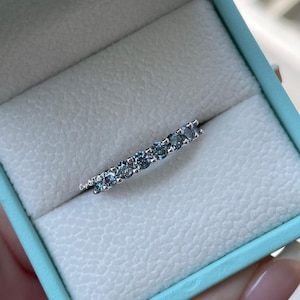 Puede incluir: Un anillo de plata con una fila de piedras preciosas azules, presentado en una caja de joyería azul claro con interior blanco. El anillo es el punto focal, mostrando las brillantes piedras preciosas. La caja está abierta, revelando el diseño del anillo.