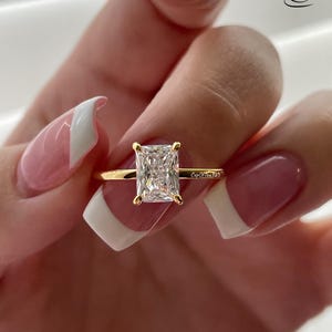 May include: A gold ring with a large, clear, square-shaped diamond. The ring is on a finger with a white and pink nail polish design. The text "CupOfTea125" is engraved on the band.
