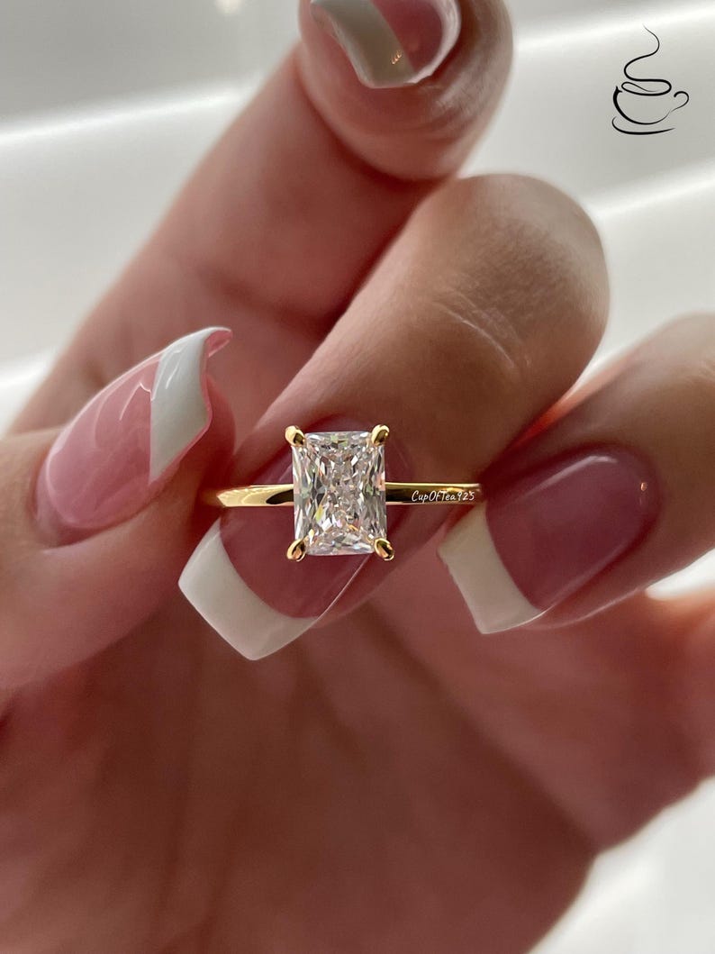 May include: A gold ring with a large, clear, square-shaped diamond. The ring is on a finger with a white and pink nail polish design. The text "CupOfTea125" is engraved on the band.