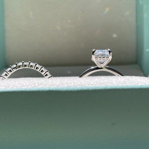 May include: A silver-toned engagement ring featuring a rectangular center stone set in a four-prong basket, accompanied by a matching curved wedding band set with multiple small, clear, rectangular-cut stones along the top half of the band.