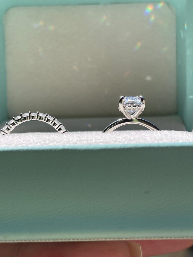 May include: A silver-toned engagement ring featuring a rectangular center stone set in a four-prong basket, accompanied by a matching curved wedding band set with multiple small, clear, rectangular-cut stones along the top half of the band.