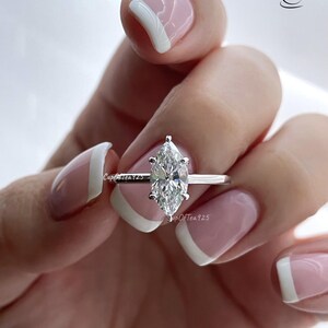 K&ouml;nnte beinhalten: Ein silberner Ring mit einem Marquise-f&ouml;rmigen Diamanten. Der Ring befindet sich an einem Finger mit wei&szlig;er French Manicure.