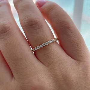 May include: A thin gold band ring featuring a continuous row of small, round, clear faceted stones set along the entire circumference. The metal has a polished yellow finish, and the stones are arranged in a prong setting to create a sparkling effect.