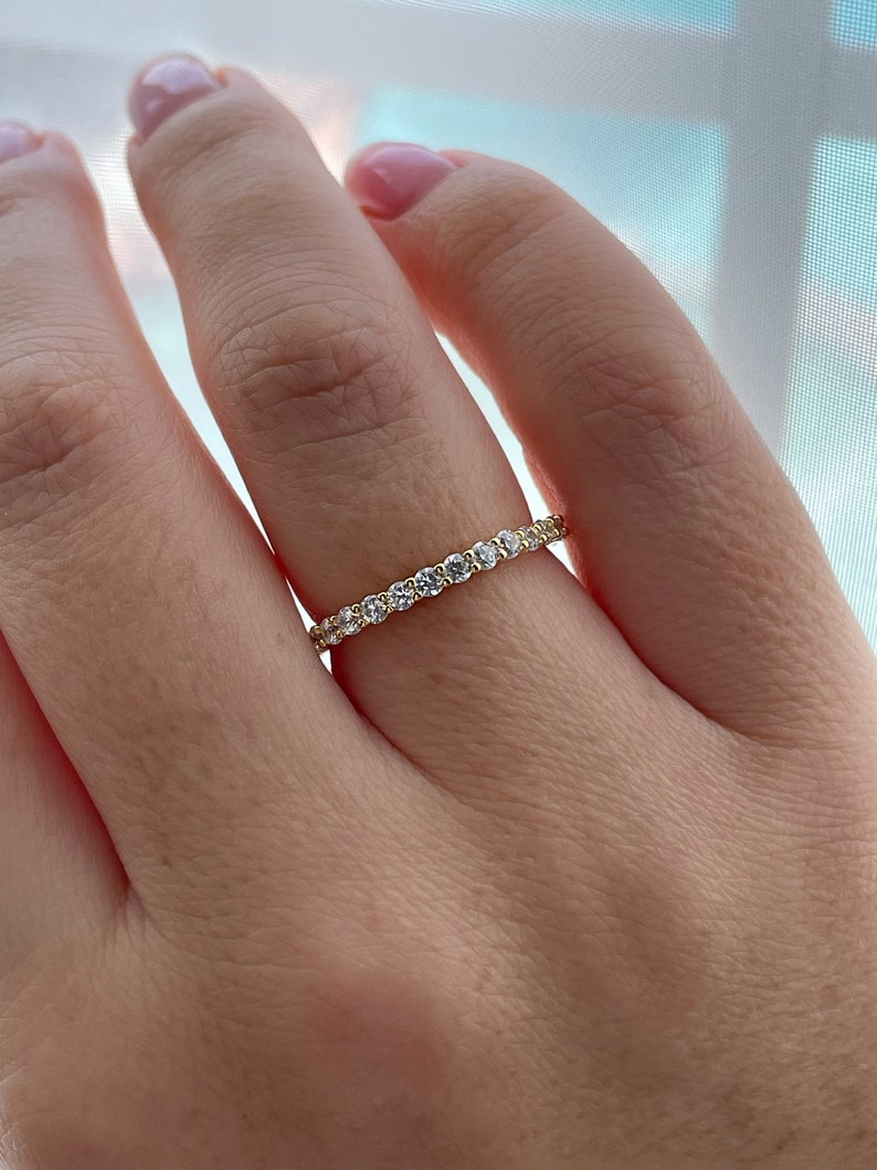 May include: A thin gold band ring featuring a continuous row of small, round, clear faceted stones set along the entire circumference. The metal has a polished yellow finish, and the stones are arranged in a prong setting to create a sparkling effect.