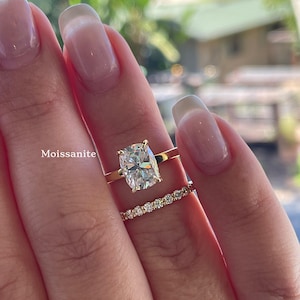 May include: A gold ring featuring a cushion-cut Moissanite center stone and a matching gold band set with a row of small round brilliant-cut stones. The word Moissanite is printed in white text next to the center stone on the gold band.