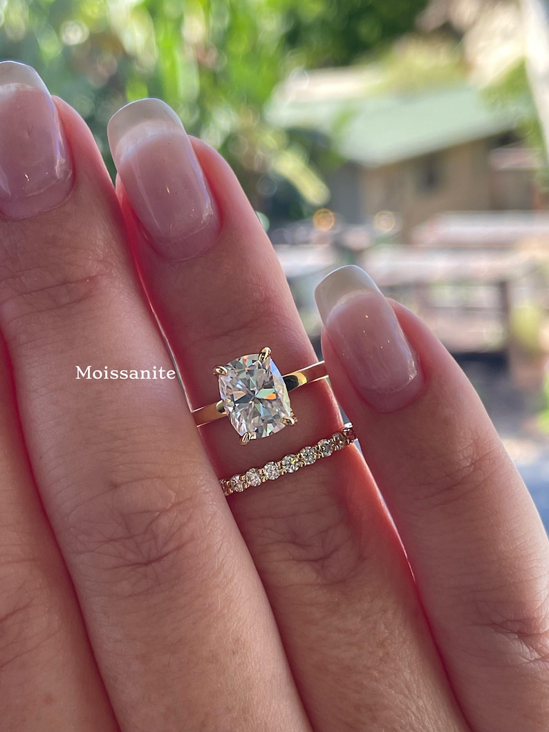 May include: A gold ring featuring a cushion-cut Moissanite center stone and a matching gold band set with a row of small round brilliant-cut stones. The word Moissanite is printed in white text next to the center stone on the gold band.