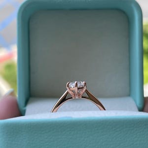 K&ouml;nnte beinhalten: Ein Verlobungsring aus Ros&eacute;gold mit einem gro&szlig;en, klaren Edelstein wird in einer hellblauen Ringschatulle pr&auml;sentiert. Der Ring hat ein klassisches, minimalistisches Design mit einer Vier-Zacken-Fassung. Die Schatulle ist ge&ouml;ffnet und zeigt den Ring.