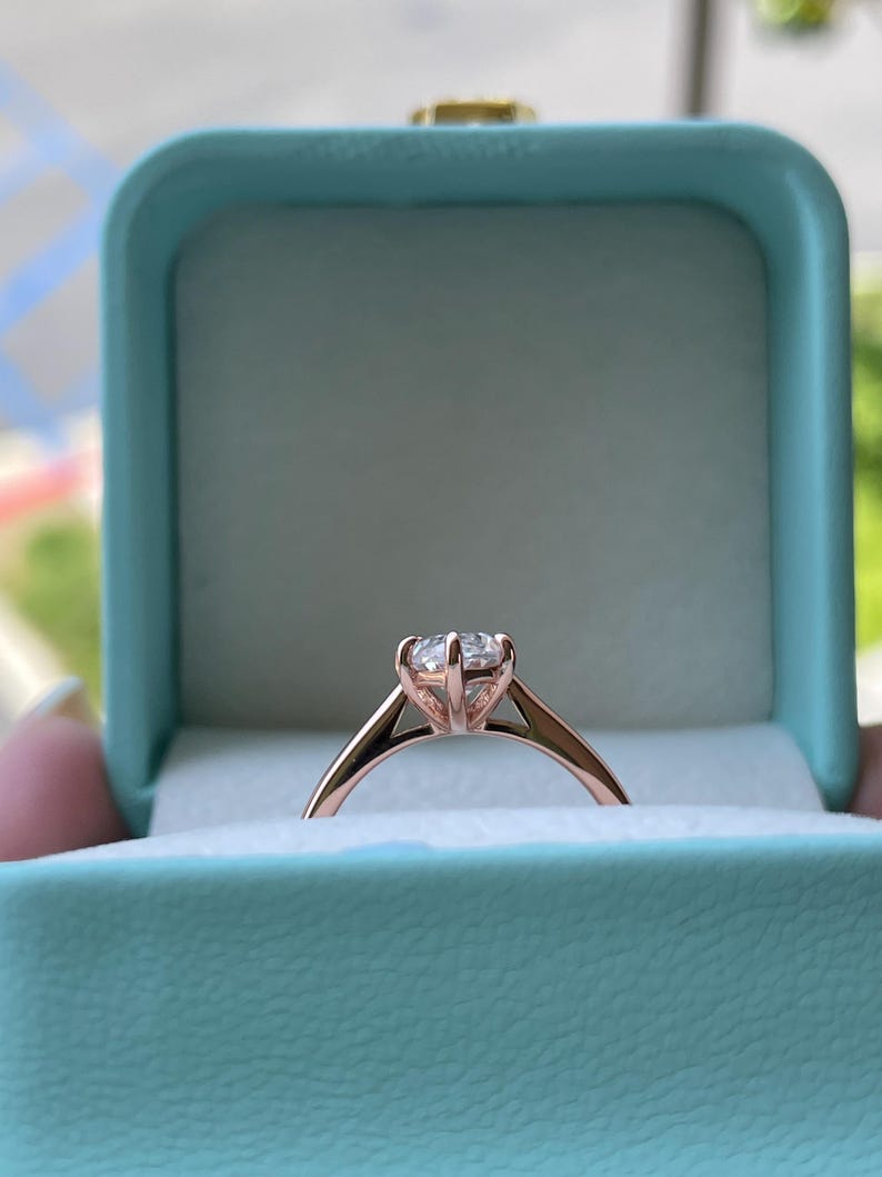 K&ouml;nnte beinhalten: Ein Verlobungsring aus Ros&eacute;gold mit einem gro&szlig;en, klaren Edelstein wird in einer hellblauen Ringschatulle pr&auml;sentiert. Der Ring hat ein klassisches, minimalistisches Design mit einer Vier-Zacken-Fassung. Die Schatulle ist ge&ouml;ffnet und zeigt den Ring.