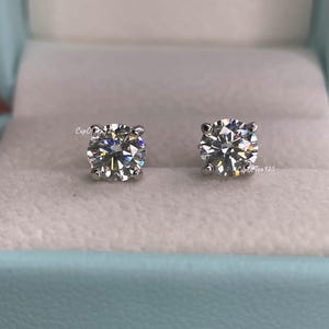 May include: A pair of silver stud earrings with a clear round gemstone in the center. The earrings are in a light blue box.