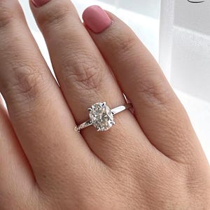 May include: A silver ring with an oval-shaped diamond. The ring is on a person's finger.