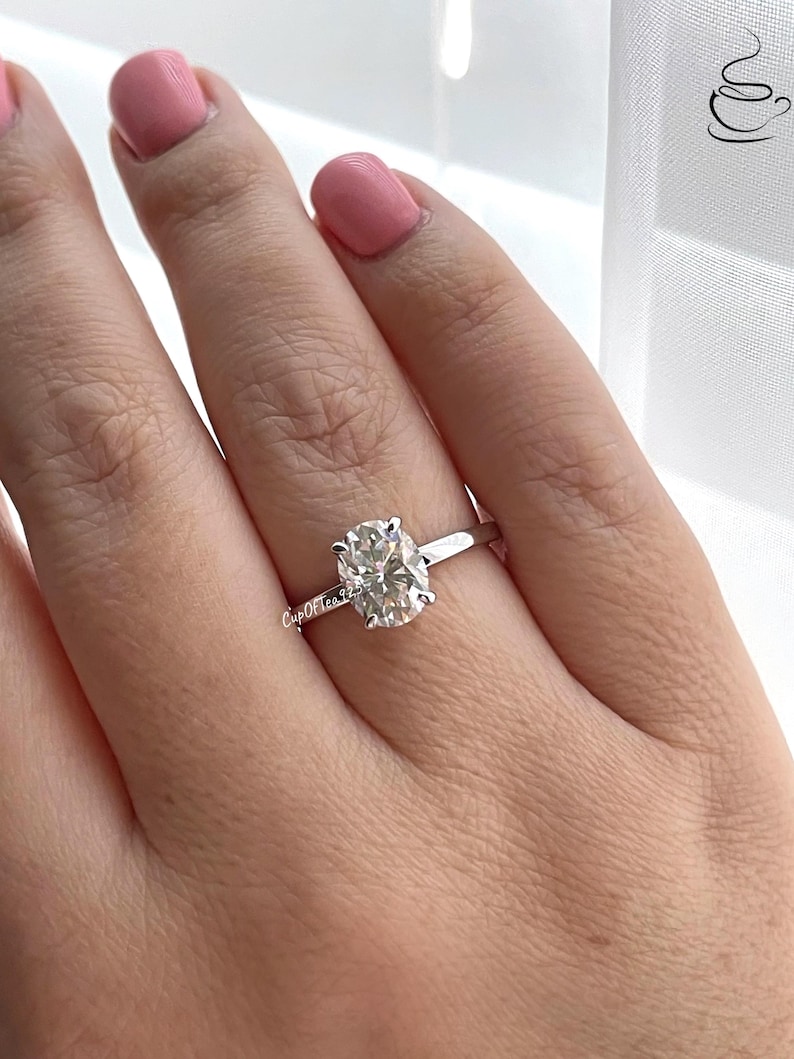 May include: A silver ring with an oval-shaped diamond. The ring is on a person's finger.
