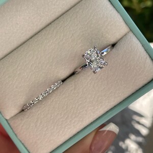 May include: A silver-toned engagement ring set featuring a cushion-cut center stone on a thin band and a matching eternity band with small round stones. The rings are displayed in a light blue box with a beige interior lining.
